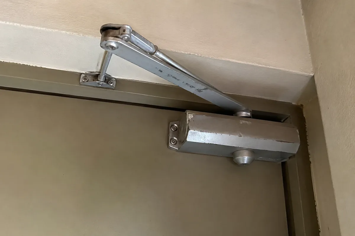 Detalle de un cierrapuertas metálico con marcas de uso instalado en la parte superior de una puerta