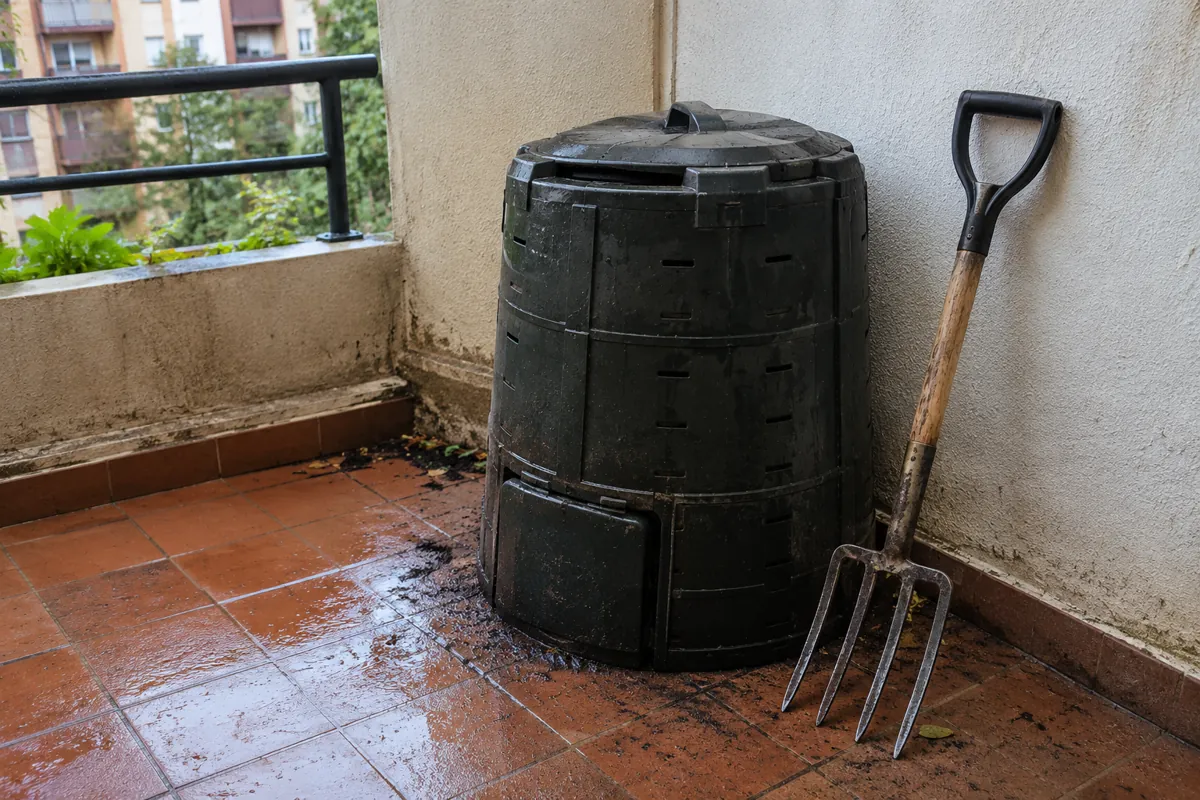 Un contenedor de compostaje cerrado y una horquilla de jardín están junto a una pared