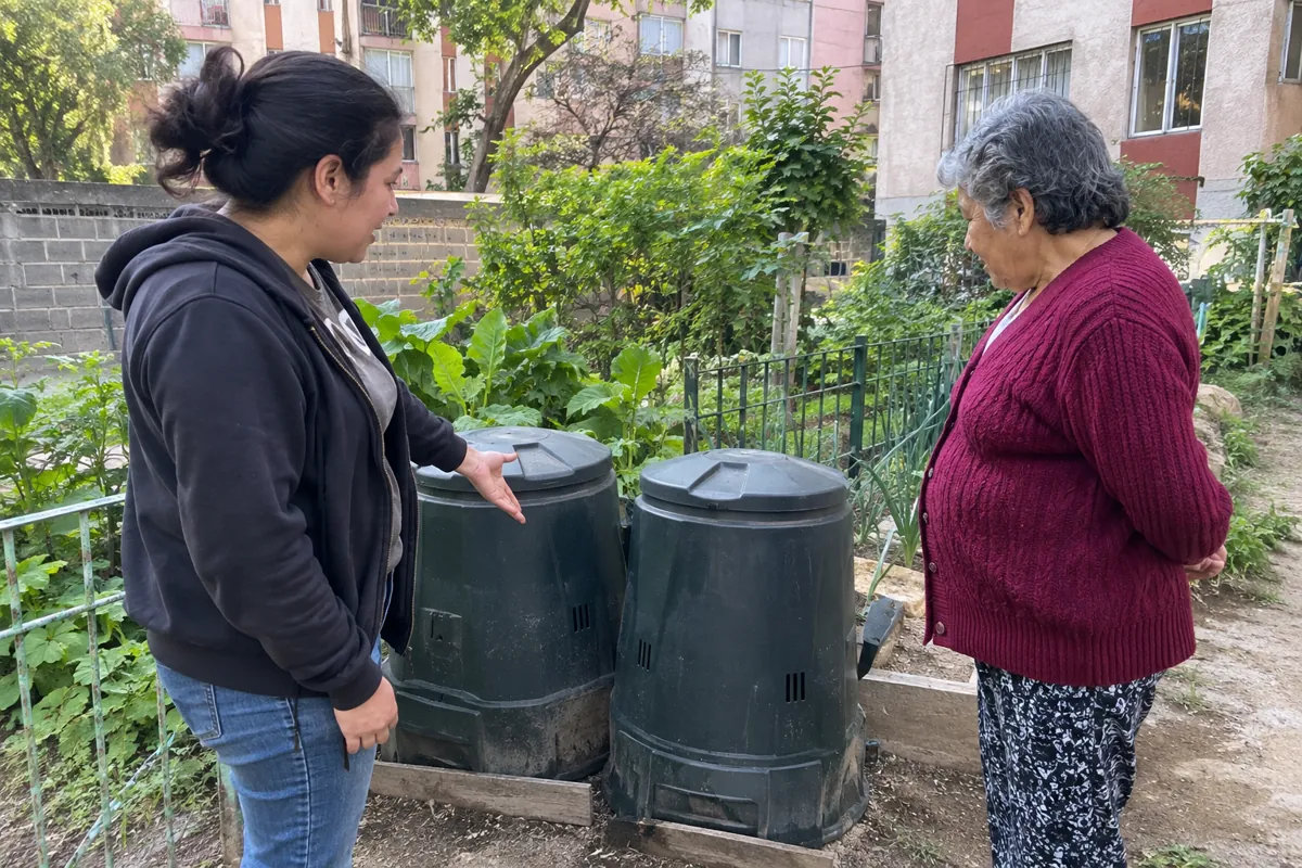 Dos mujeres conversan junto a dos composteras cerradas en un huerto