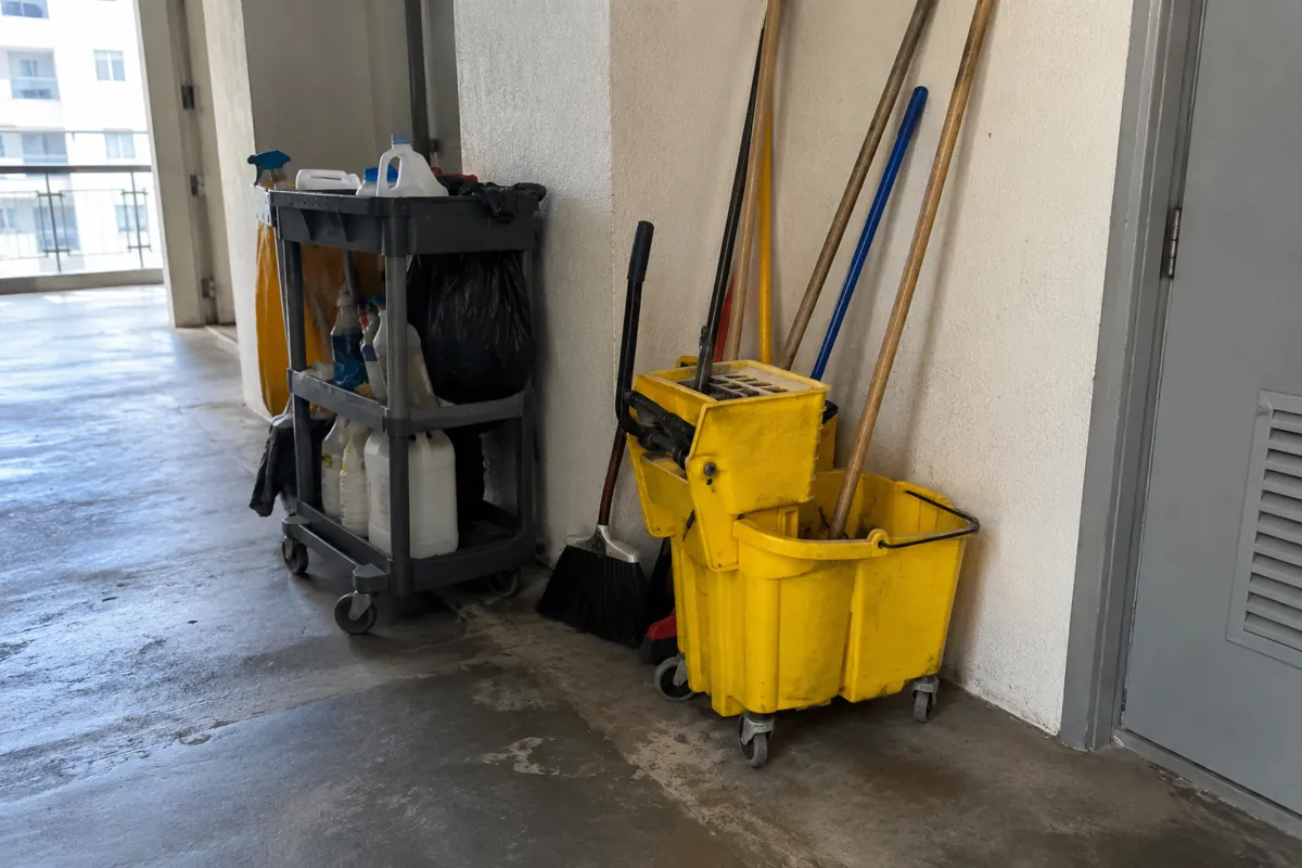 Carrito, cubeta con exprimidor, escobas y trapeadores guardados junto a una pared