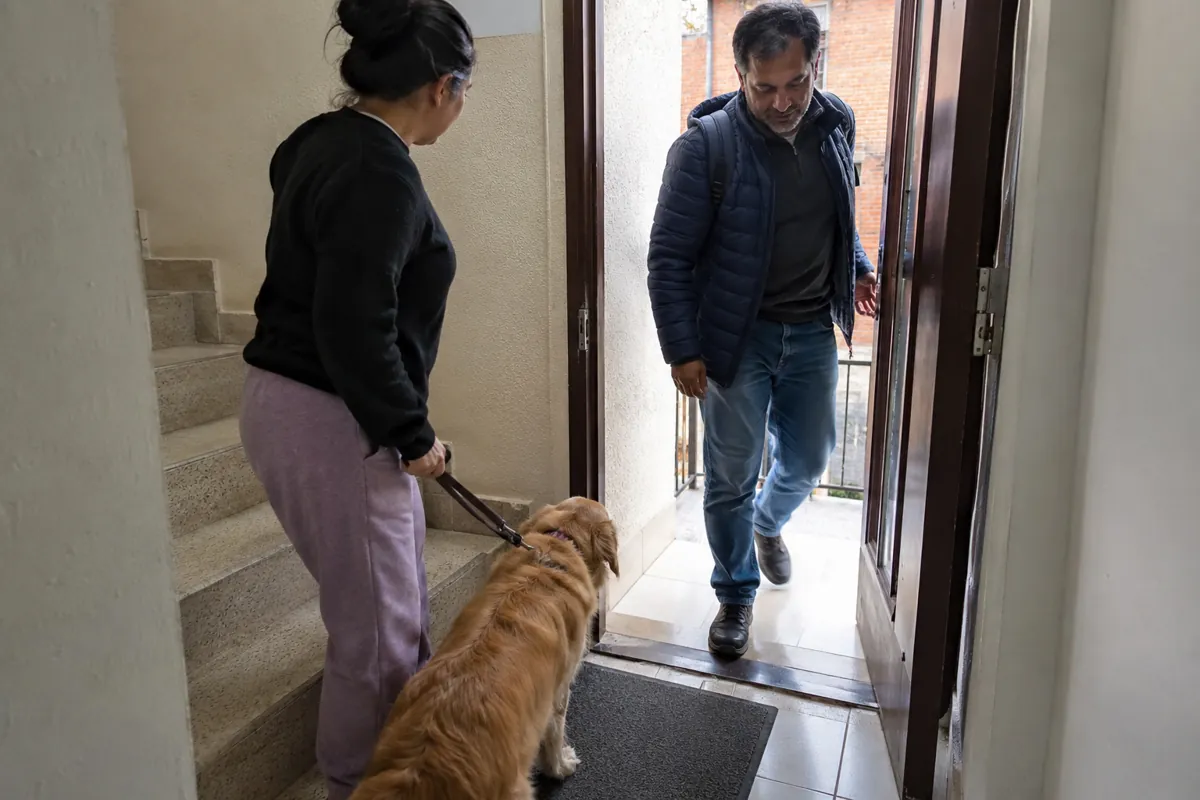 Una mujer sostiene la correa de un perro junto a la escalera mientras un hombre entra por la puerta