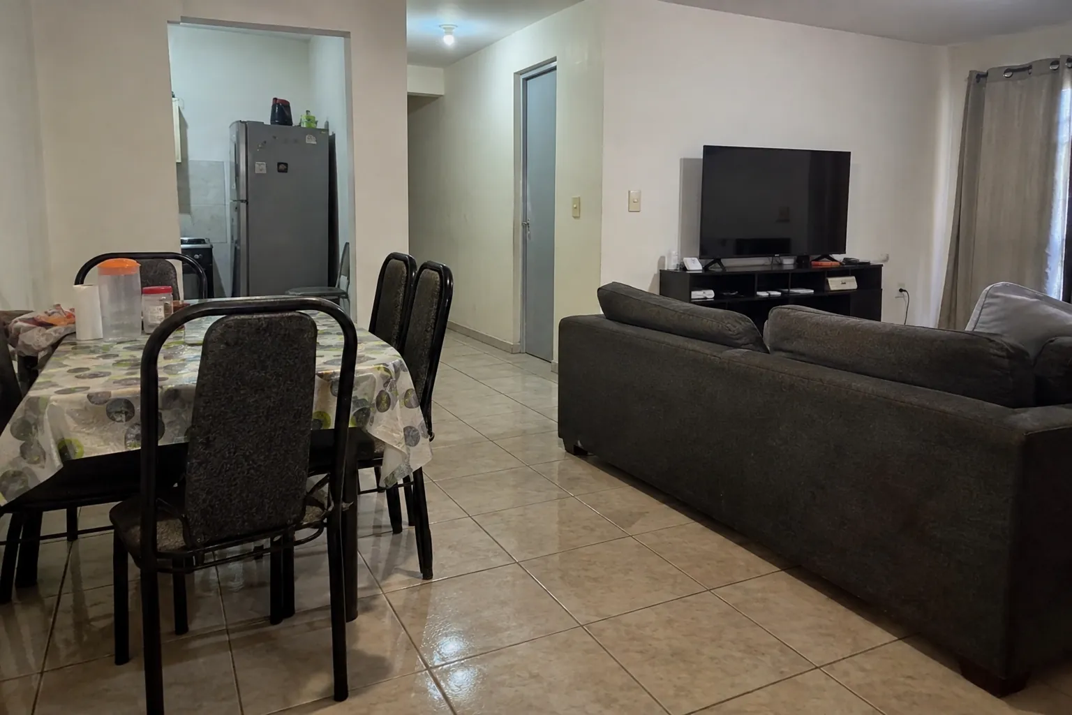 Salón vacío con mesa y sillas en un lado y sofá frente a una televisión apagada en el otro