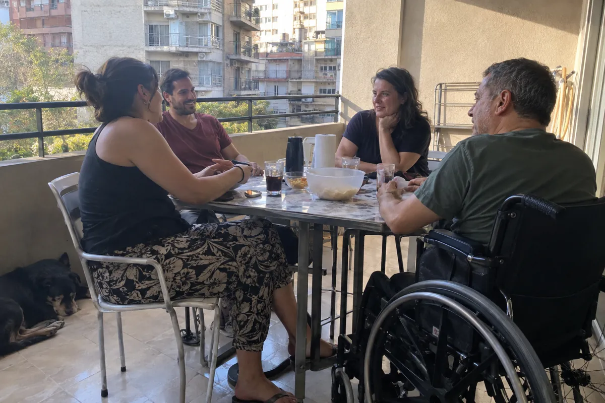 Cuatro personas adultas conversan alrededor de una mesa en un balcón, una de ellas en silla de ruedas