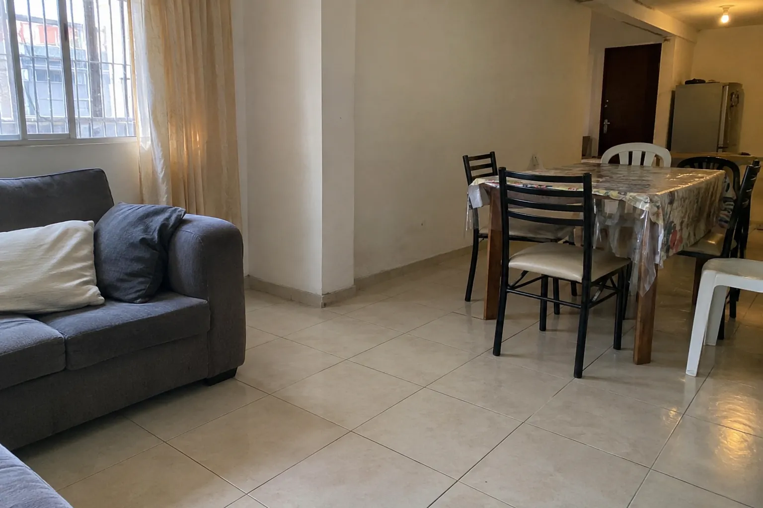 Salón vacío con sofá junto a una ventana y mesa rodeada de sillas distintas, con espacio libre entre ambos