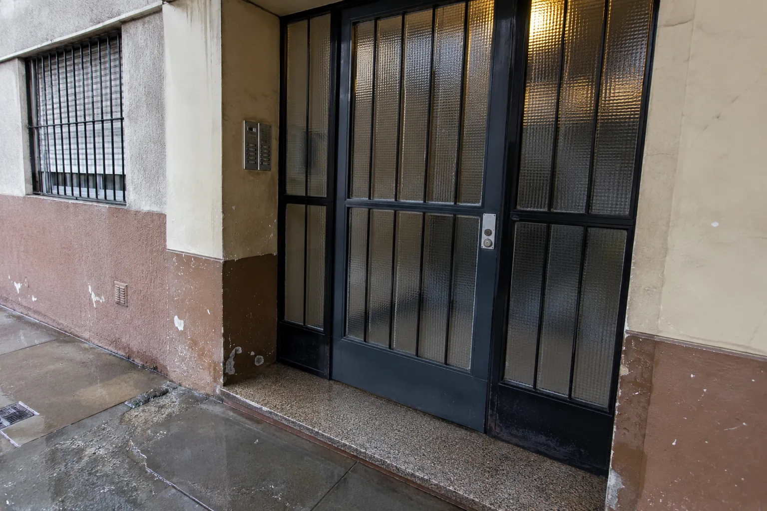 Puerta de entrada cerrada de metal y vidrio junto a un intercomunicador en una fachada con pintura desgastada